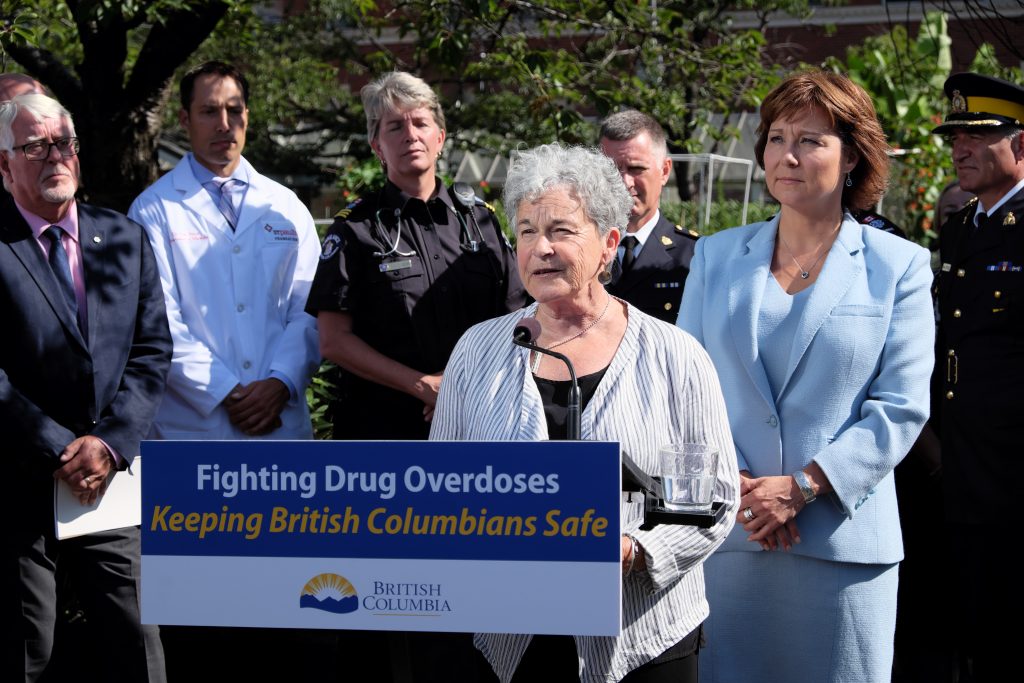 Woman stands at podium which reads Fighting Drug Overdoses Keeping British Columbians Safe. Behind her a number of people, including two police officers, a doctor, and a paramedic, stand somberly.