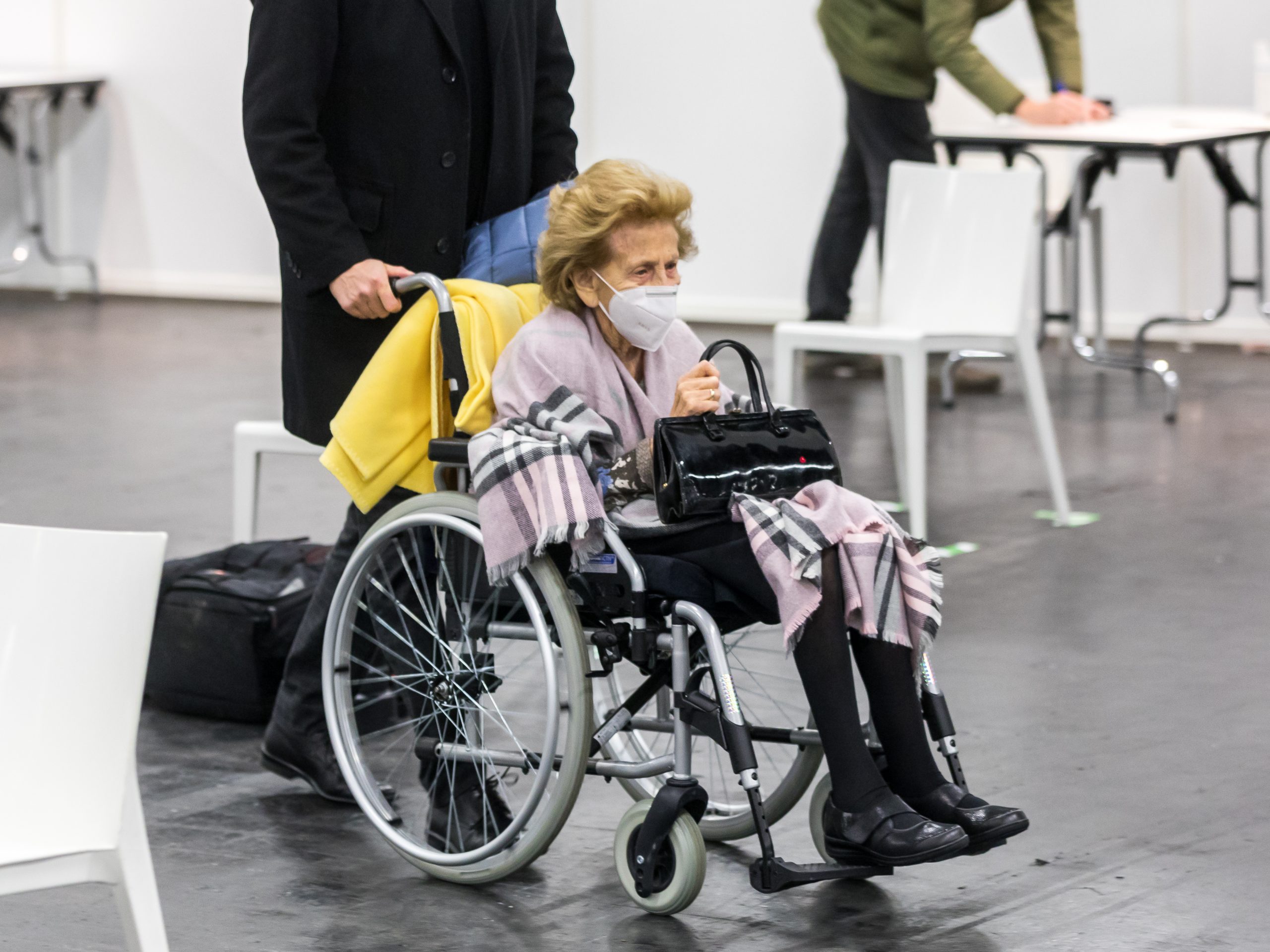 Photo showing an elderly woman, wearing a mask being pushed in a wheelchair