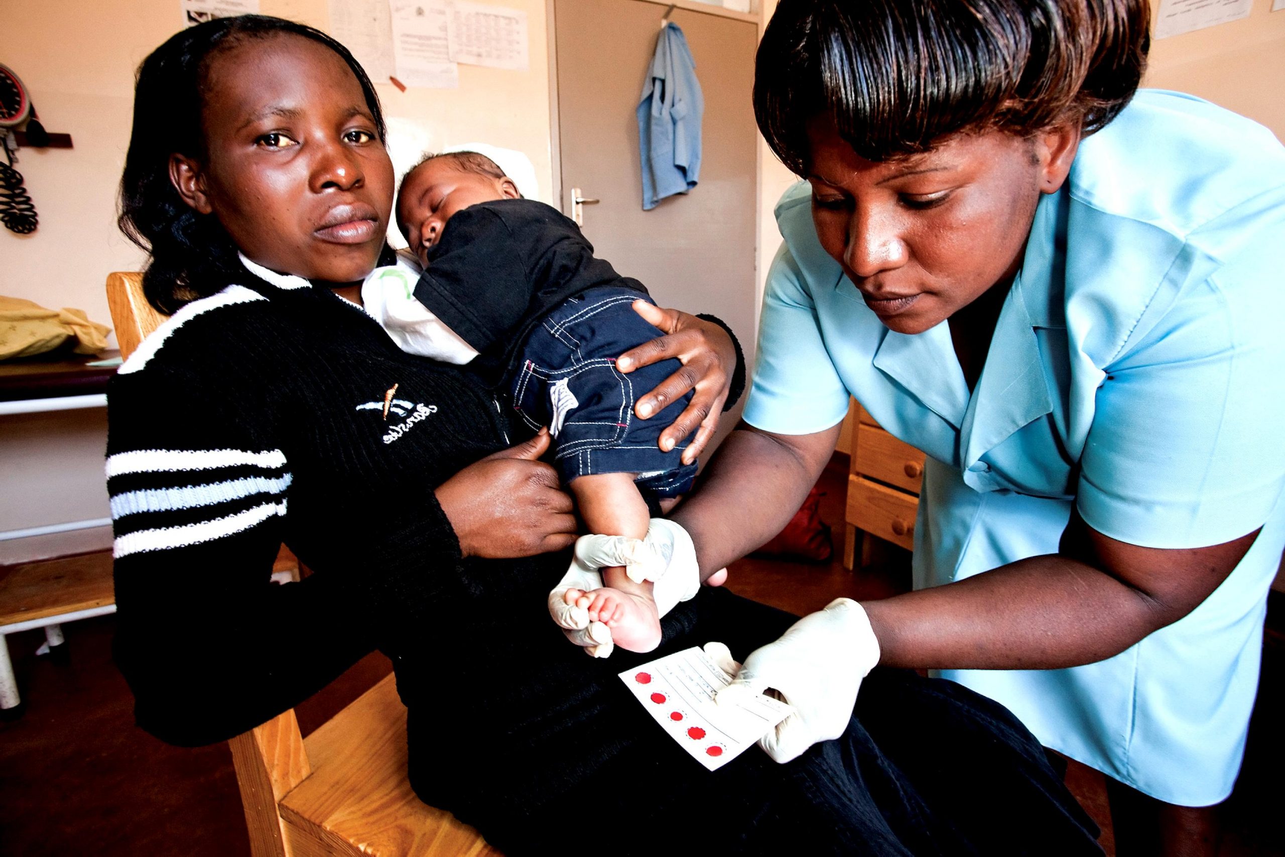 Photo showing a seated mother holding an infant while a nurse performs health screenings on the infant