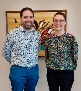 Jérôme Melançon has short beard and long hair, tied back; Emily Grafton has long hair, tied back, and glasses. They are standing side by side, and both have their hands behind their backs. Both are wearing flowery shirts.