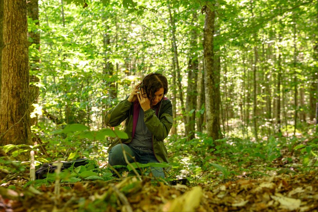 Image of a person with brown curly hair in a striped shirt, jeans and a jacket crouching in the shade of a forest wearing headphones, looking at the ground, and listening attentively