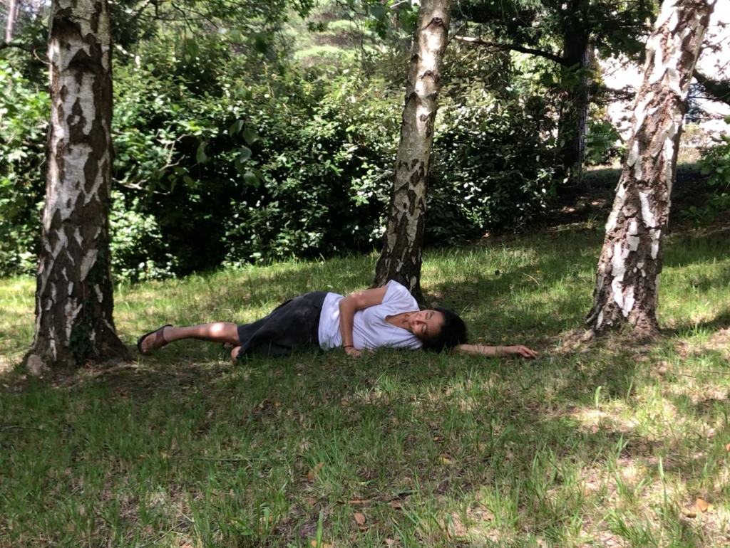 The image shows a woman wearing a white t-shirt and a gray skirt lying on the ground between three trees. Only the lower part of the trunks of the trees are visible, and the ground is covered with green grass. In the background, there is a big bush with green leaves.