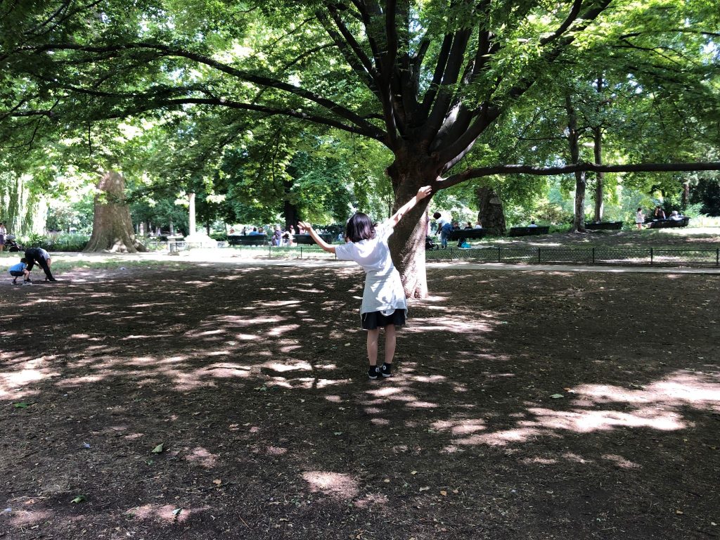 A woman wearing a white t-shirt and a gray skirt stands beneath a large tree with her arms outstretched in an upward direction. The image was taken in a park environment, and it features other people and trees with green leaves.