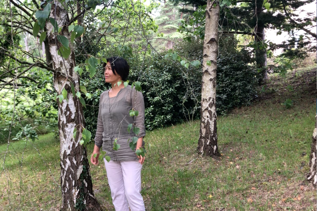 In the image, a woman stands beneath a tree on her right side, wearing a gray shirt and white pants. There are some trees and bushes, and on the ground, there is grass along with orange/brown leaves.