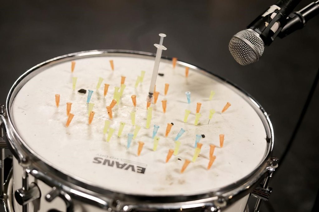 Image documentation of Lorelei d'Andriole's "The Body is an Instrument and My Instrument is a Brick." The photo is of a snare drum with many needle hubs sticking out, with a need and syringe stuck in the middle of the drum head. A microphone is seen in the corner of the photograph, implying that the sounds of the snare drums injection are amplified.