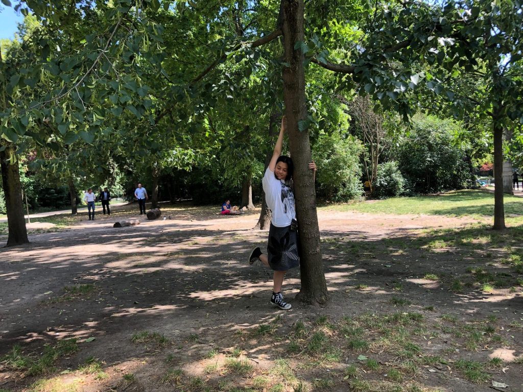 In the image, a woman wearing a white t-shirt and a gray skirt stands beneath a tree, holding onto the tree with her hands and lifting one of her feet. The image was taken in a park environment, and it features other people and trees with green leaves.