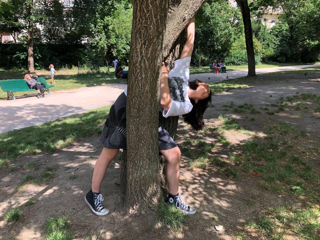 A woman wearing a white t-shirt and a gray skirt grabs a tree trunk and bends her body to her left side in the space between the double trunk of the tree. The image was taken in a park and also features other people, trees, and a green bench.