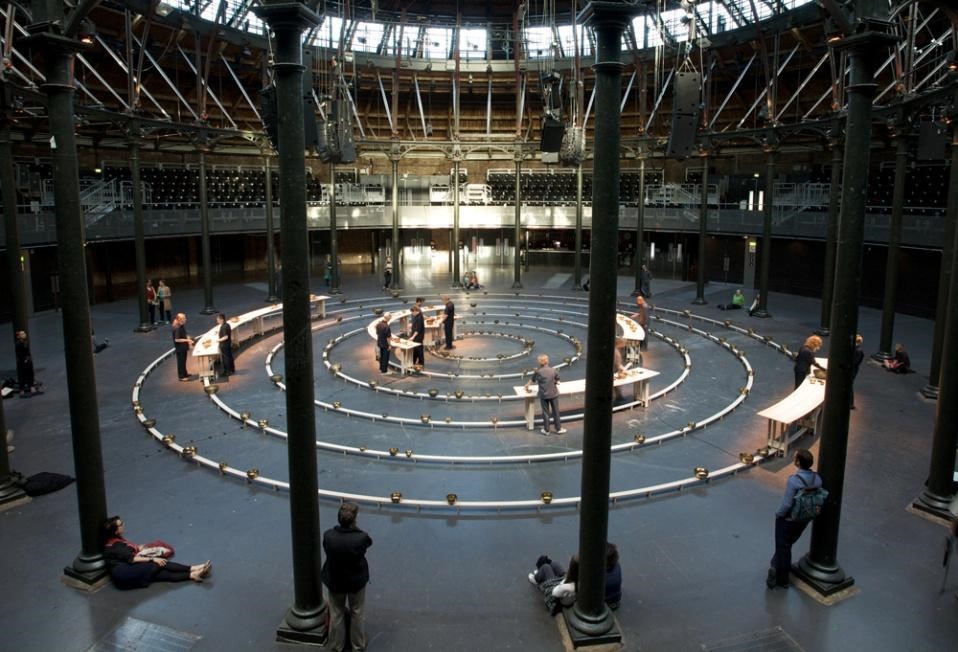 A photo of musicians playing singing bowls in The Roundhouse in London in 2009. The musicians are arranged in a circular pattern to match the graphic score.