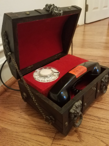 Two photos of a small wood chest with imitation Spanish baroque-style ornamental, latches, hinges, etc. Photo on the right shows the chest open to show it is lined with red fabric has been constructed to hold a fully functional rotary dial analog telephone.