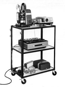 AV equipment cart of the sort used in classrooms, containing a film projector, slide projector, and open-reel audio tape recorder.