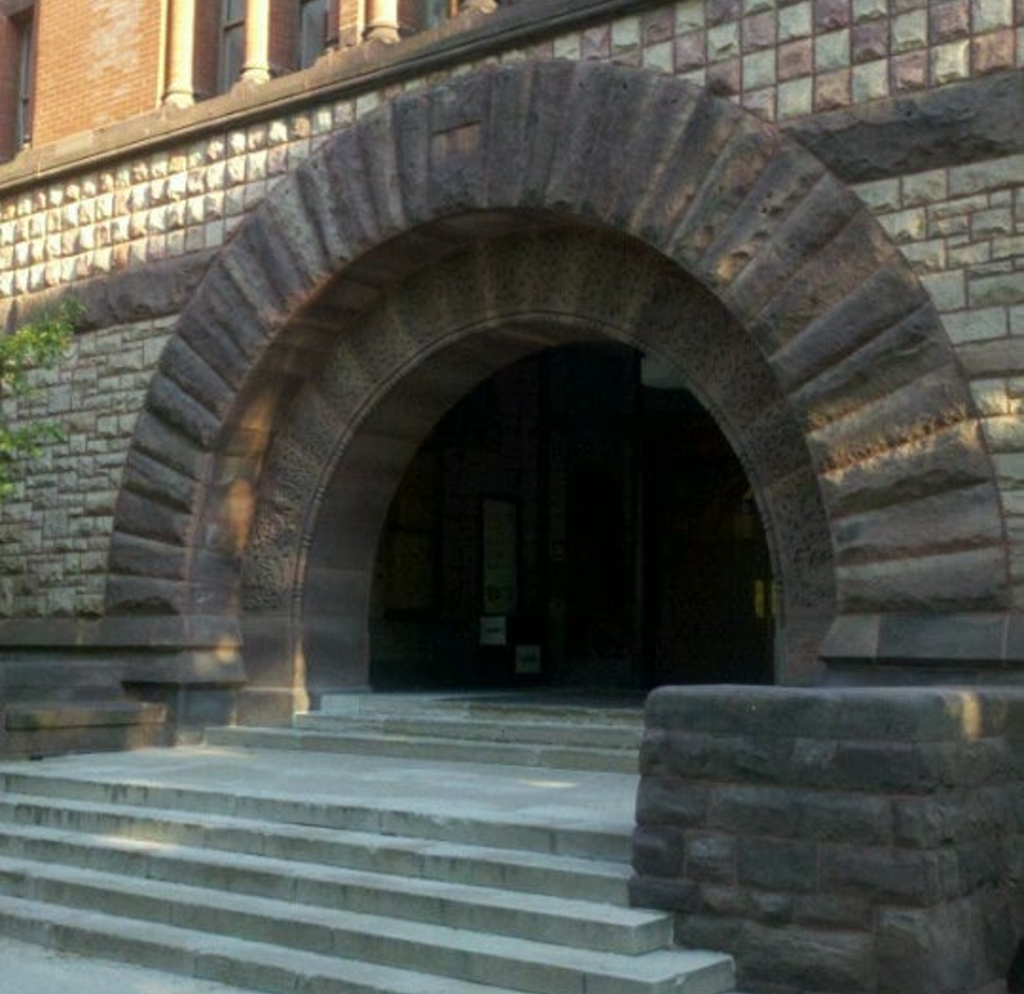 Facade of a building with a wide but low set of stairs leading to a porch and wide heavy stone archway. The curves of this arch resemble the arches traversing the negative space in the preceding drawing.