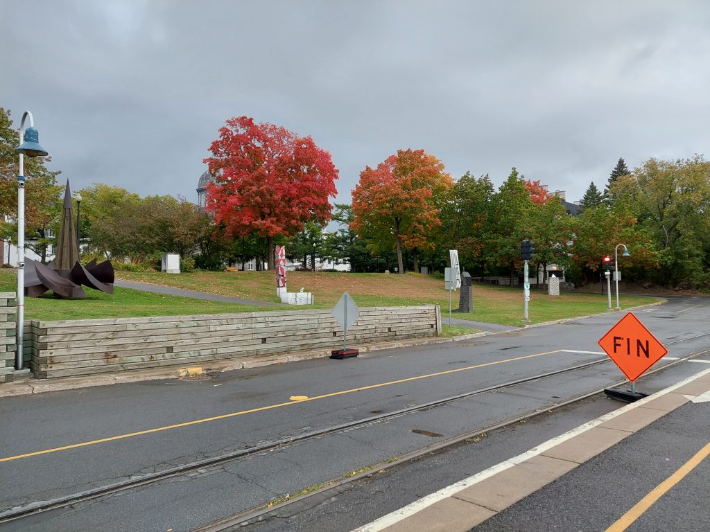 A street, a small retaining wall, a public park, trees with red, orange, and green foliage, cloudy skies. On the right, there is an orange construction signs that says "FIN."